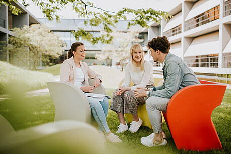 Drei junge Menschen sitzen im Campusgarten und unterhalten sich.