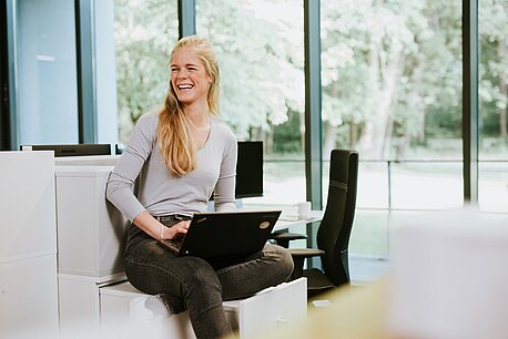 Eine Frau sitzt mit einem Laptop auf dem Schoß auf einem Schreibtisch.